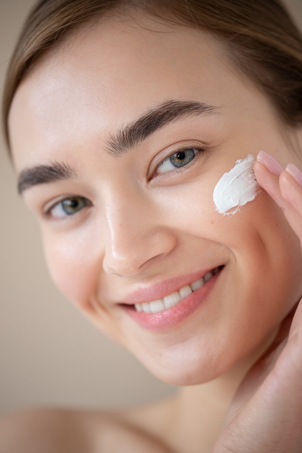 A portrait of a beautiful woman with clear skin applying moisturizer to her face, smiling gently.