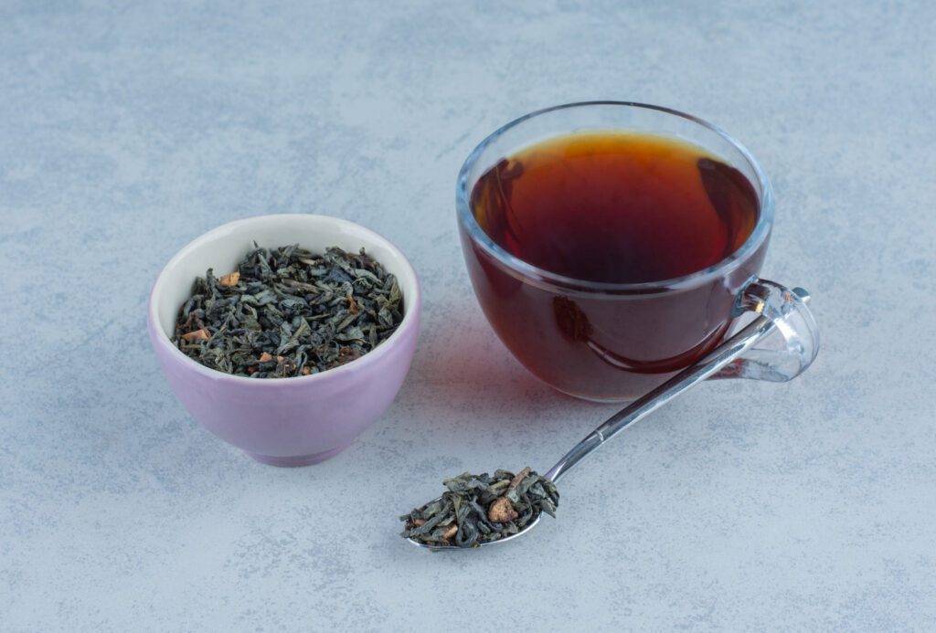 Bowl of dried black tea leaves and a cup of freshly brewed tea with a spoon on a marble surface, a natural ingredient in effective tan removal remedies and skin care.



